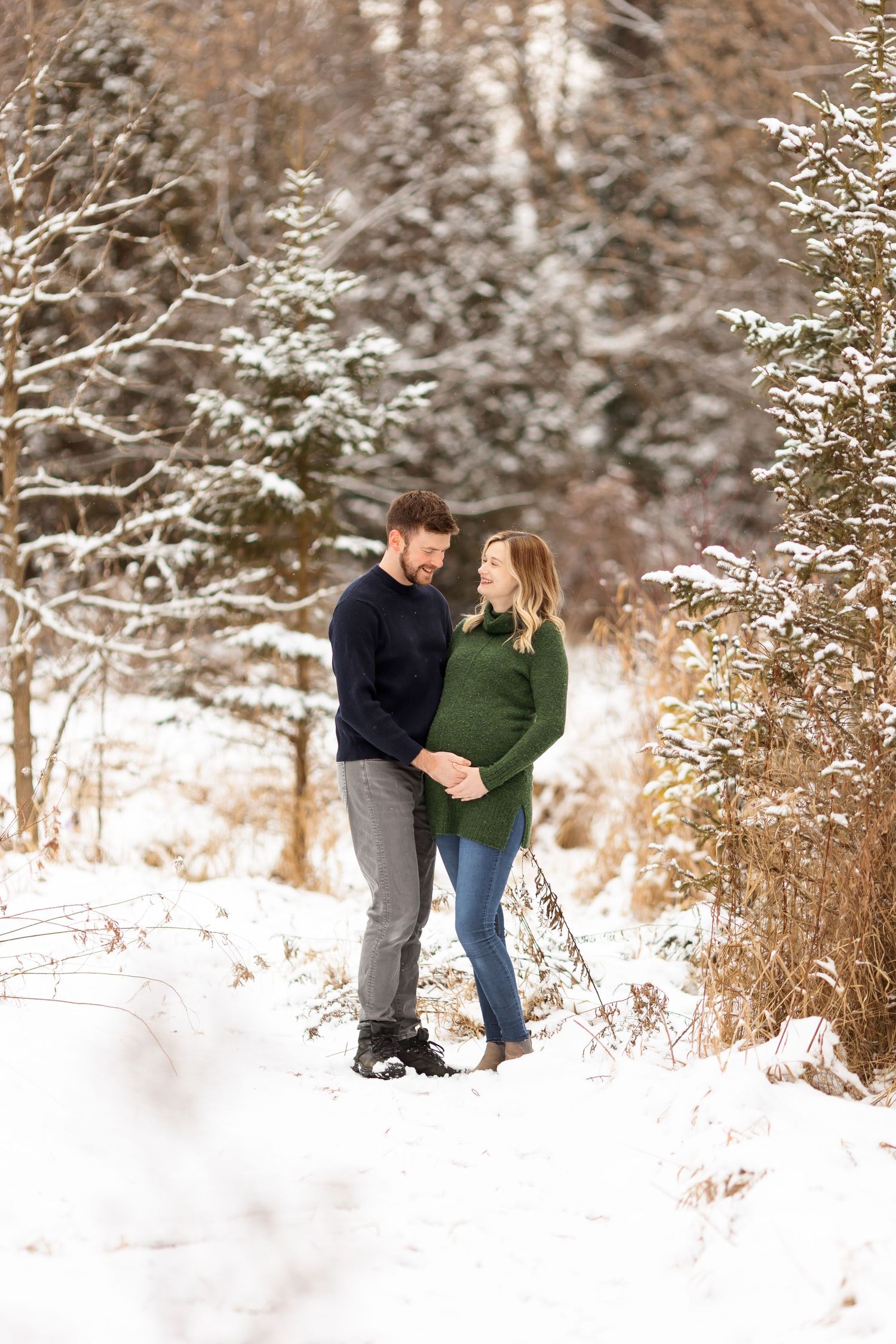A couple embraces during a winter maternity photoshoot in a snowy forest setting.