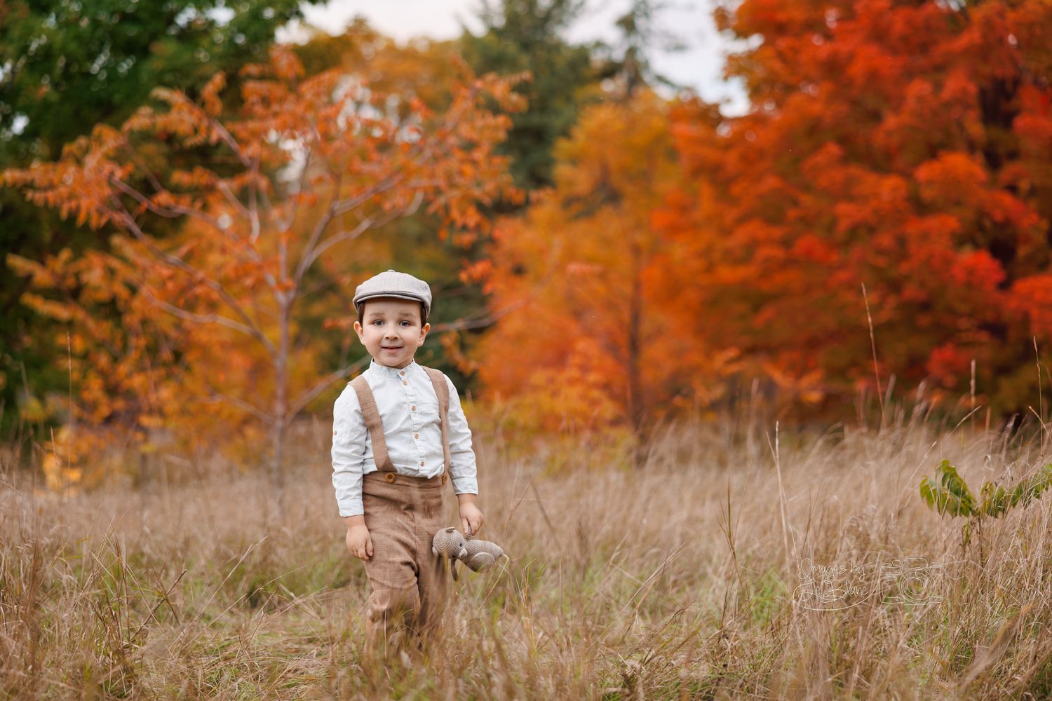 Small child wearing vintage-style outfit standing in autumn field with vibrant orange foliage in background.