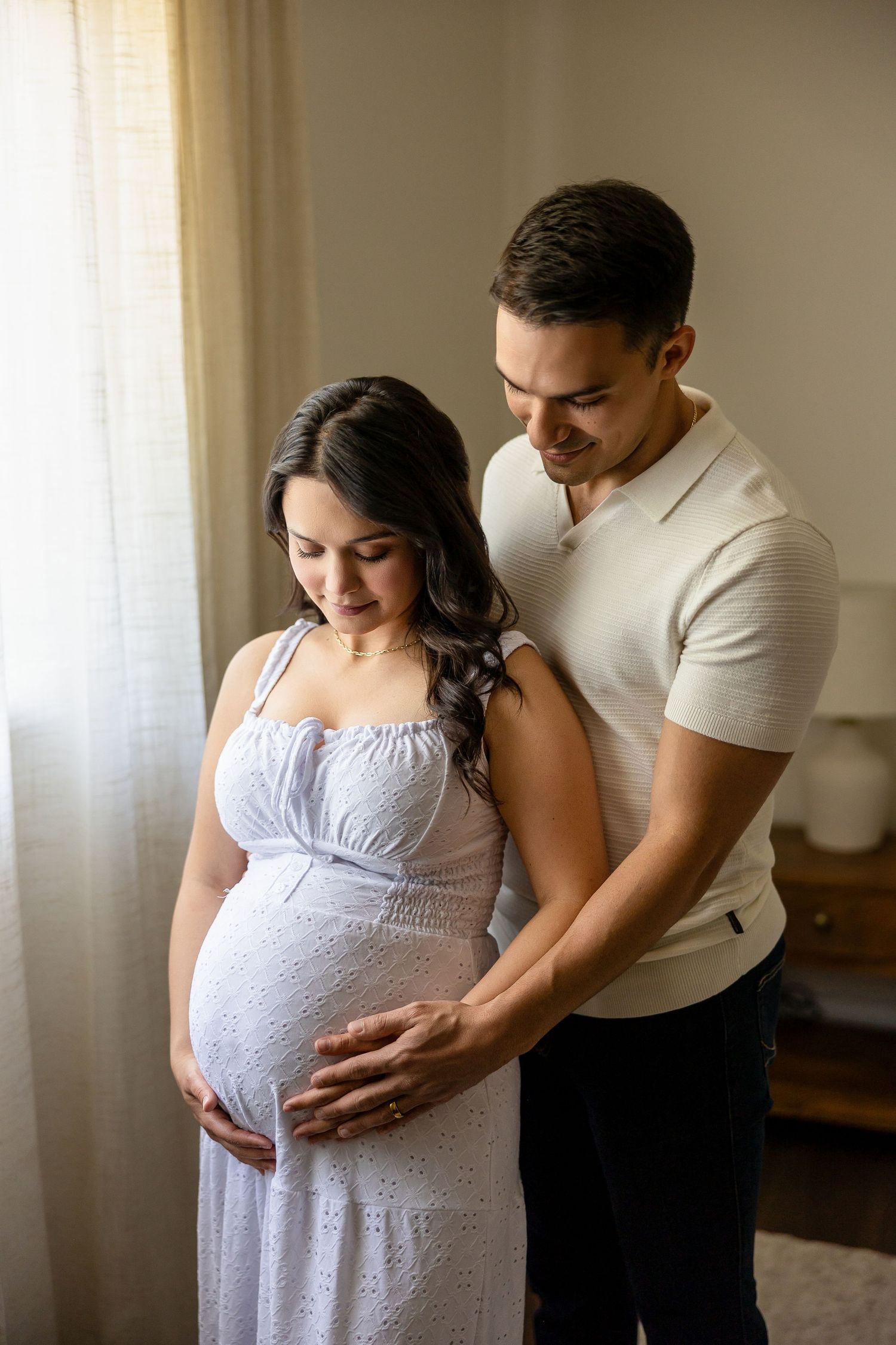 Expecting couple shares tender moment by window as they lovingly embrace growing baby bump in natural light.