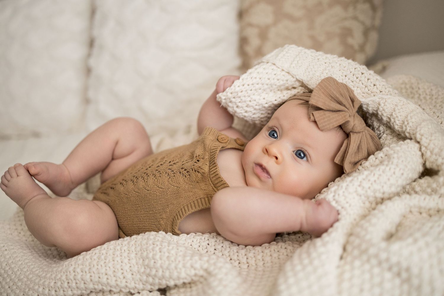 Adorable baby girl in knit romper and bow headband lying on cozy cream blanket with blue eyes.