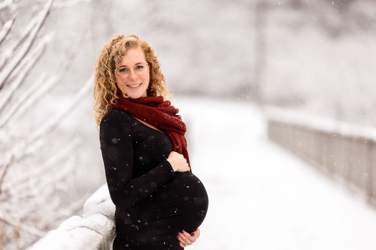 Winter maternity portrait with woman in black coat and red scarf amid falling snow.