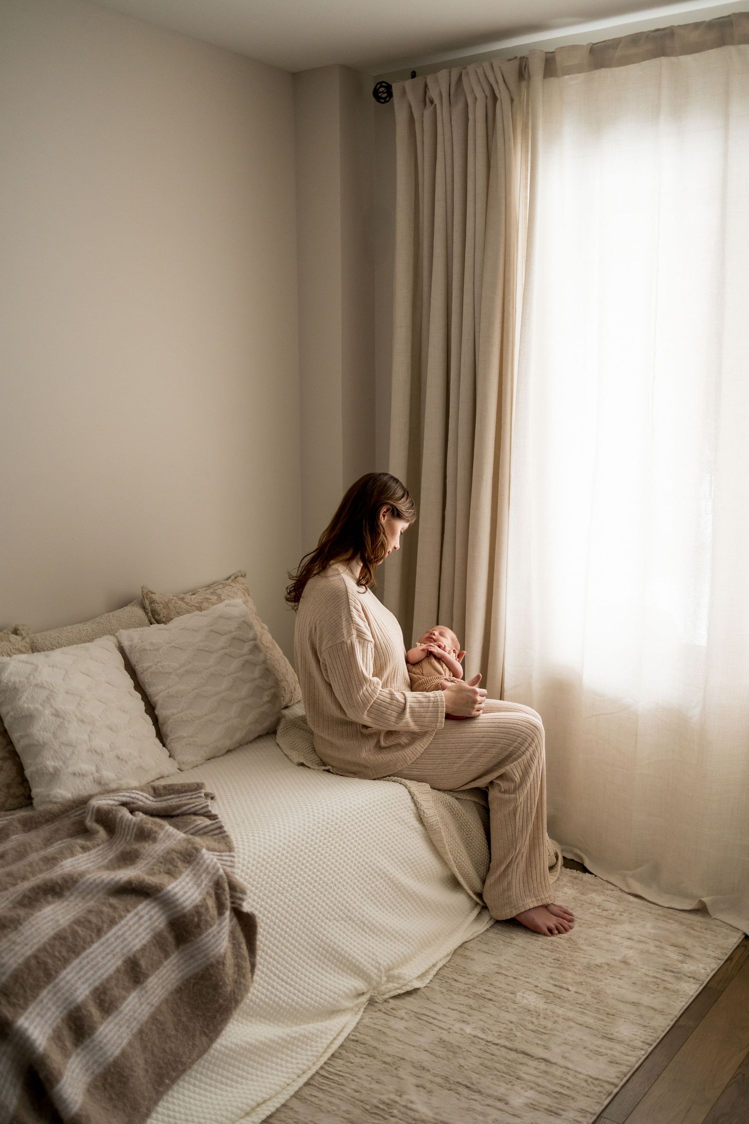 A person sits on a bed by a window in a minimalist bedroom with neutral beige decor and soft natural lighting.