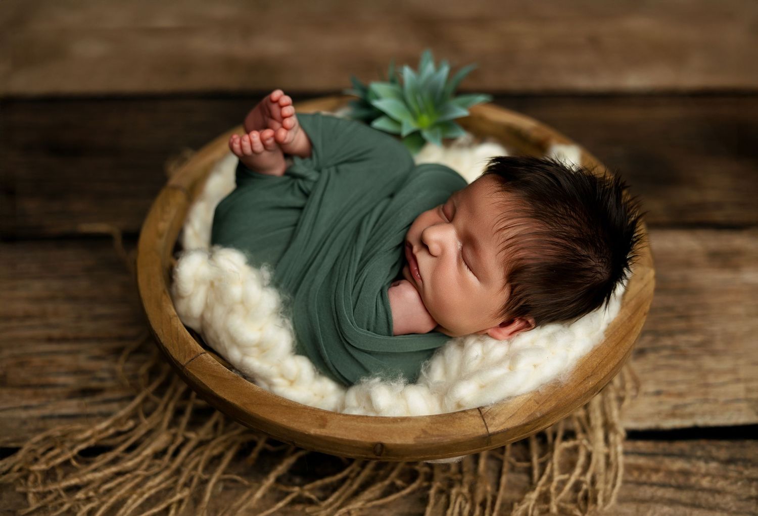 A newborn baby in green sleeps peacefully in a wooden bowl filled with soft white fabric on rustic wooden planks.