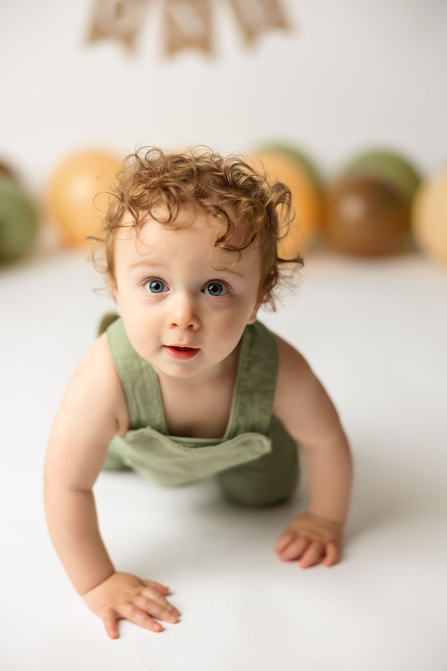 Toddler in green overalls crawling on white surface with blurred decorations in background.