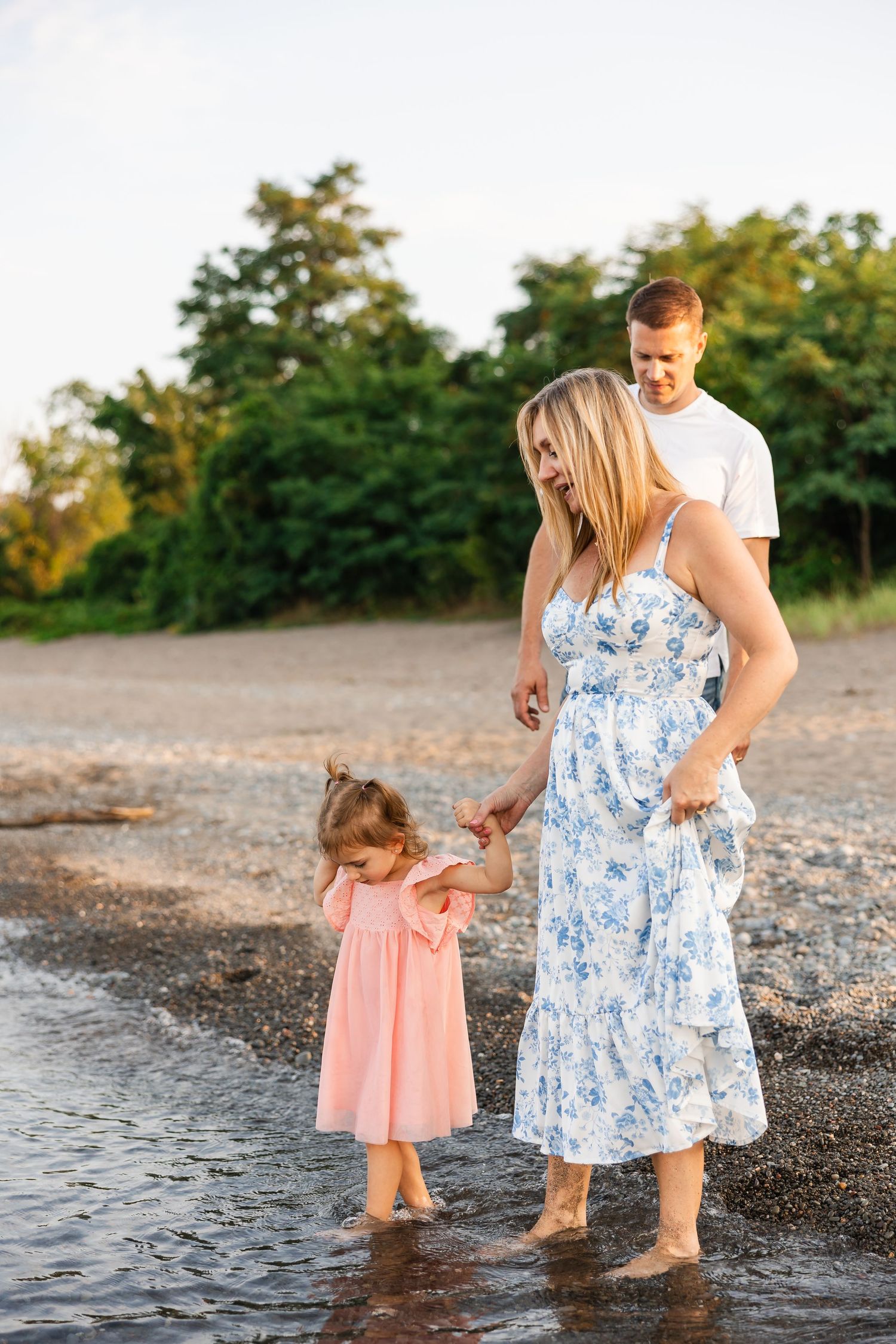 A family in summer attire stands at the water's edge during golden hour, with greenery in the background.