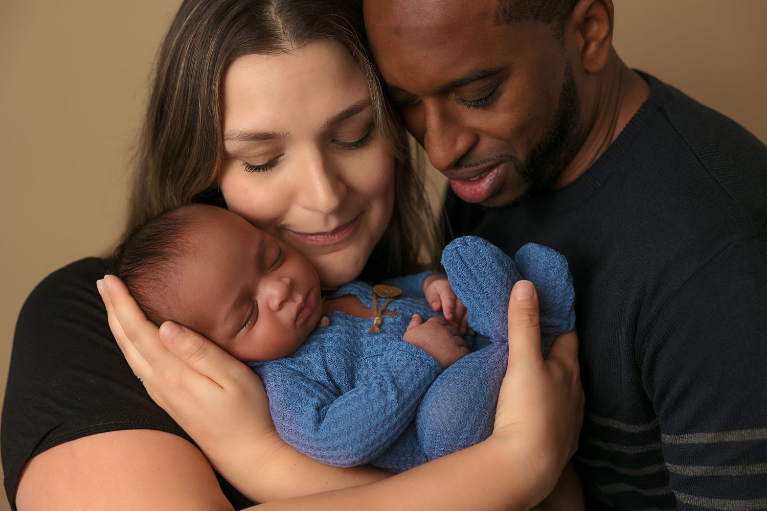 Parents sharing loving moment with sleeping baby in blue outfit against brown backdrop.