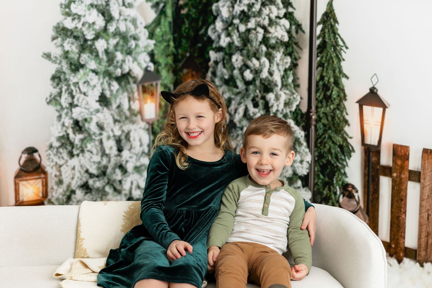 Two children sit together on white couch with snowy Christmas trees and lanterns in background.