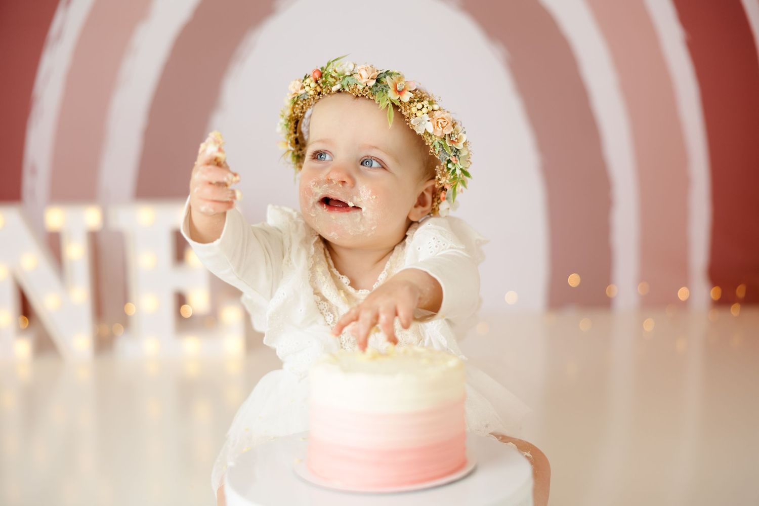 First birthday photoshoot featuring white dress and floral crown against pink rainbow background.