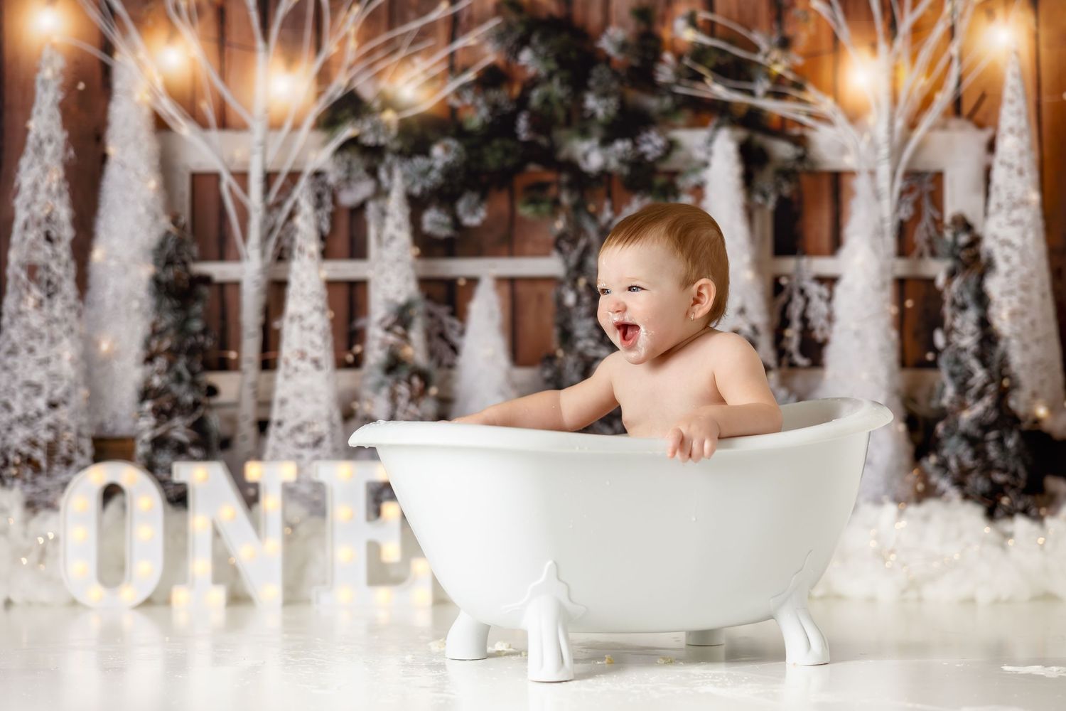 Baby sitting in white bathtub prop with illuminated ONE sign and snowy winter forest backdrop.