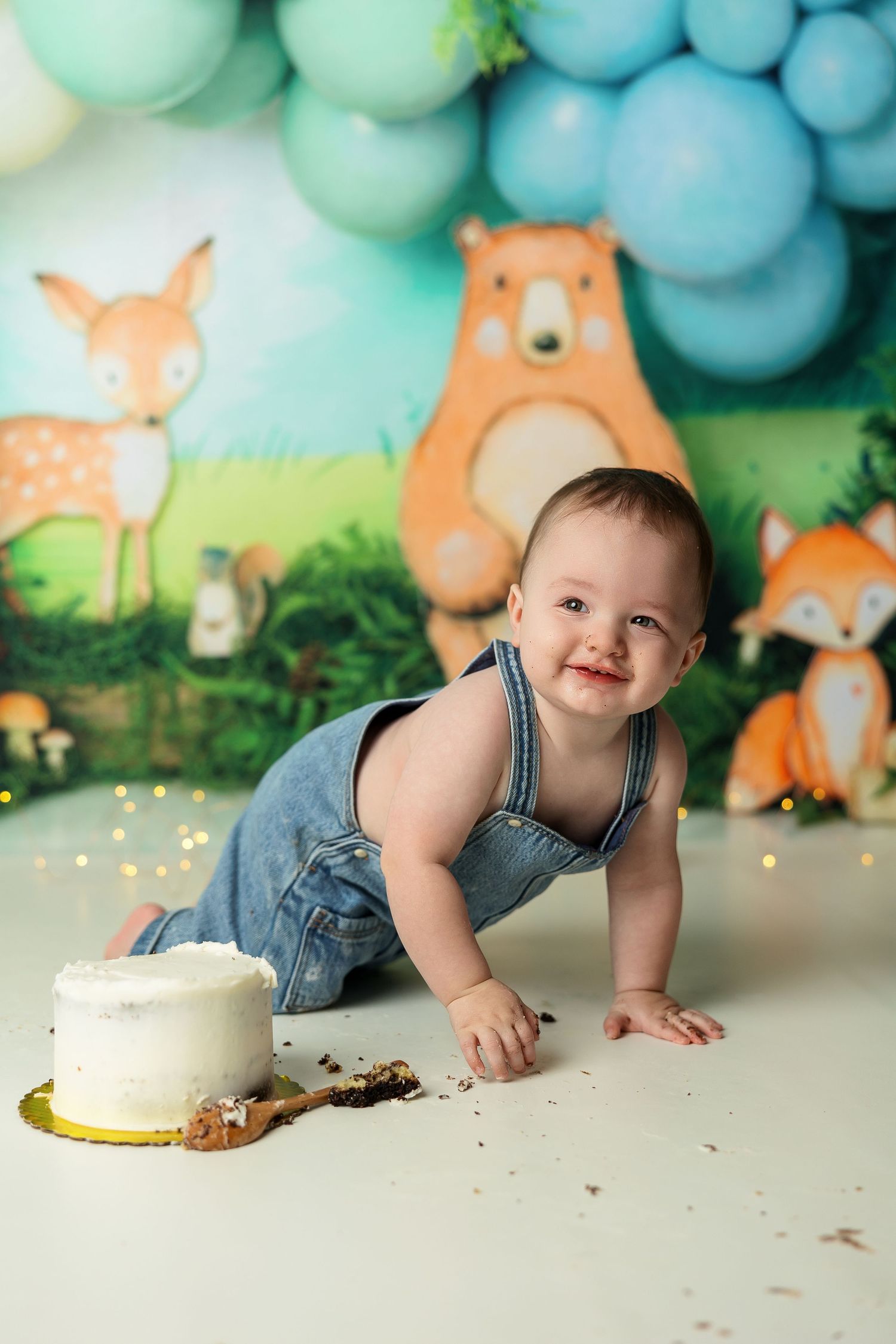 Baby in denim overalls smiling during a cake smash photoshoot with woodland animal backdrop and blue balloons.