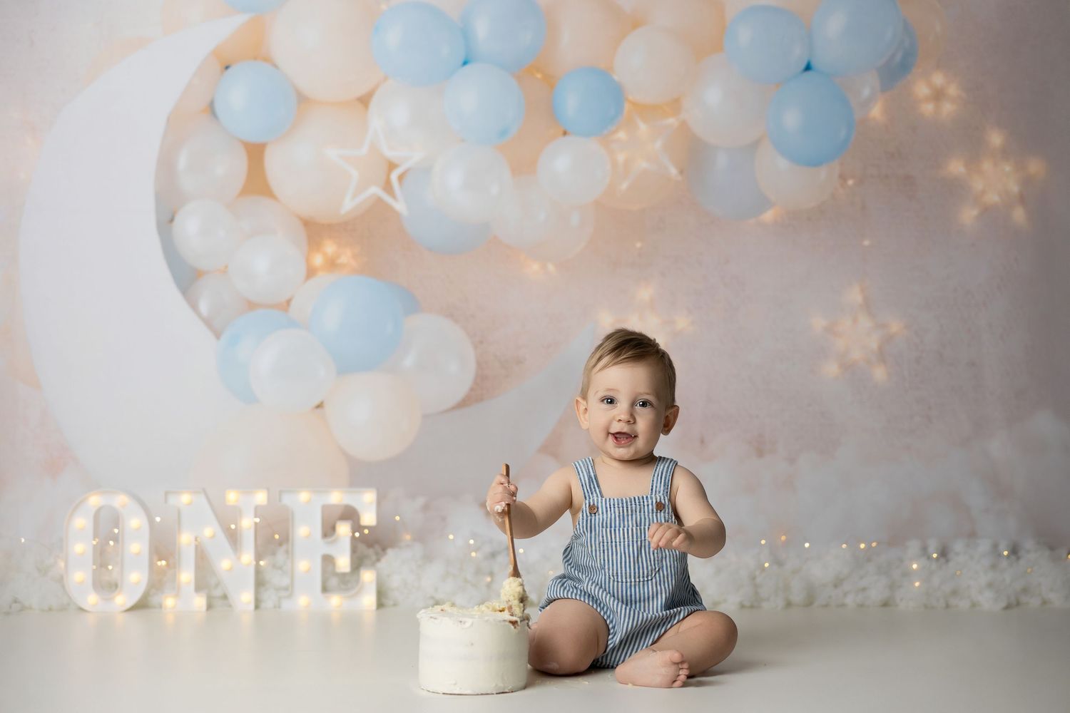A cake smash photo session with balloons, lights, and a white cake against a peach and blue backdrop.
