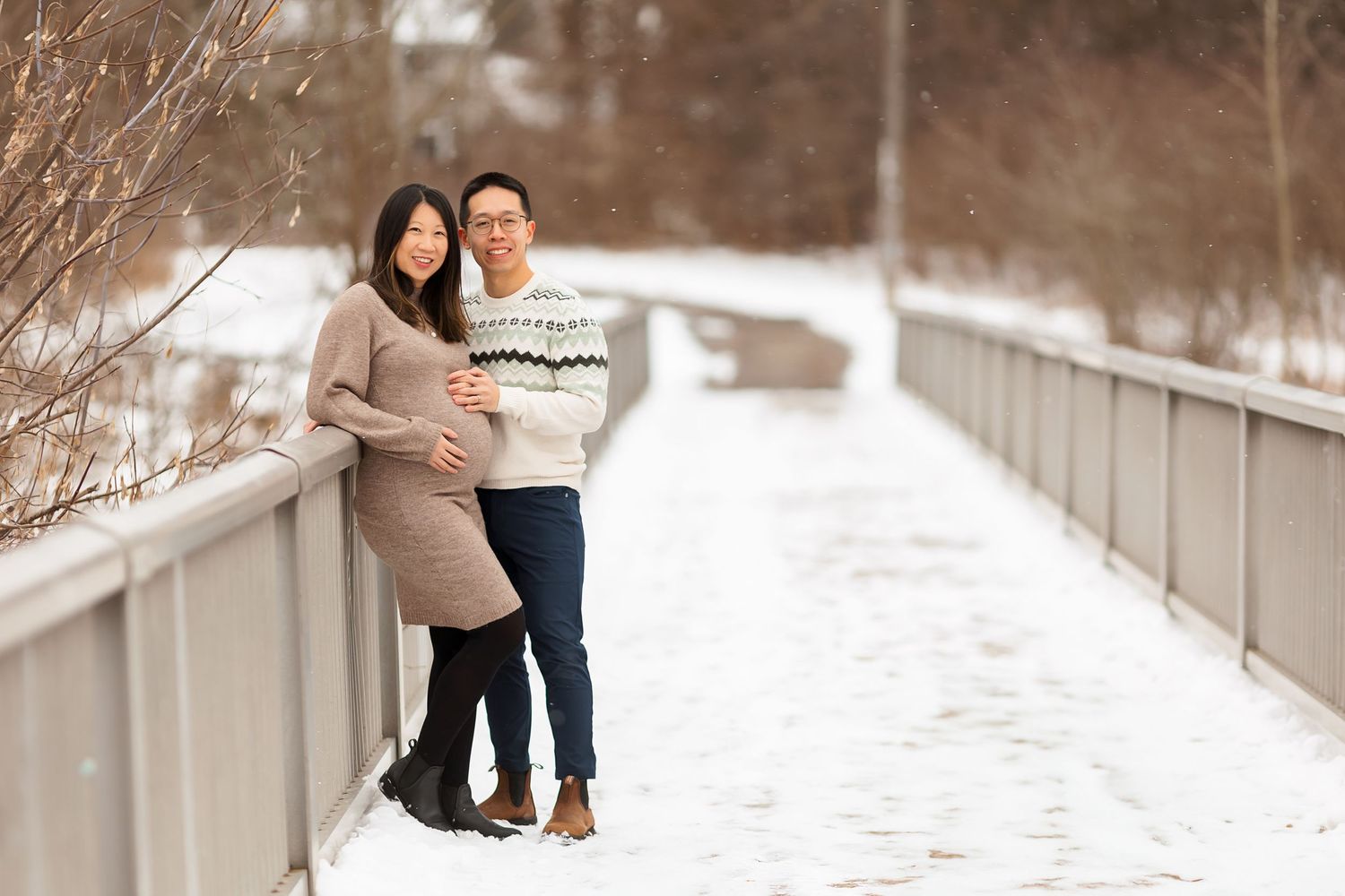 Winter maternity photos on a snow-covered bridge with evergreen trees in the background.