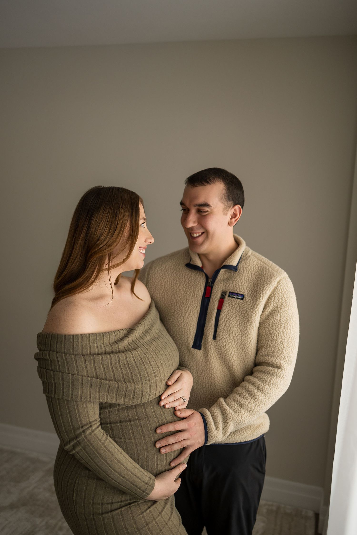 Candid maternity session captures couple sharing joyful moments in cream sweater and olive dress.
