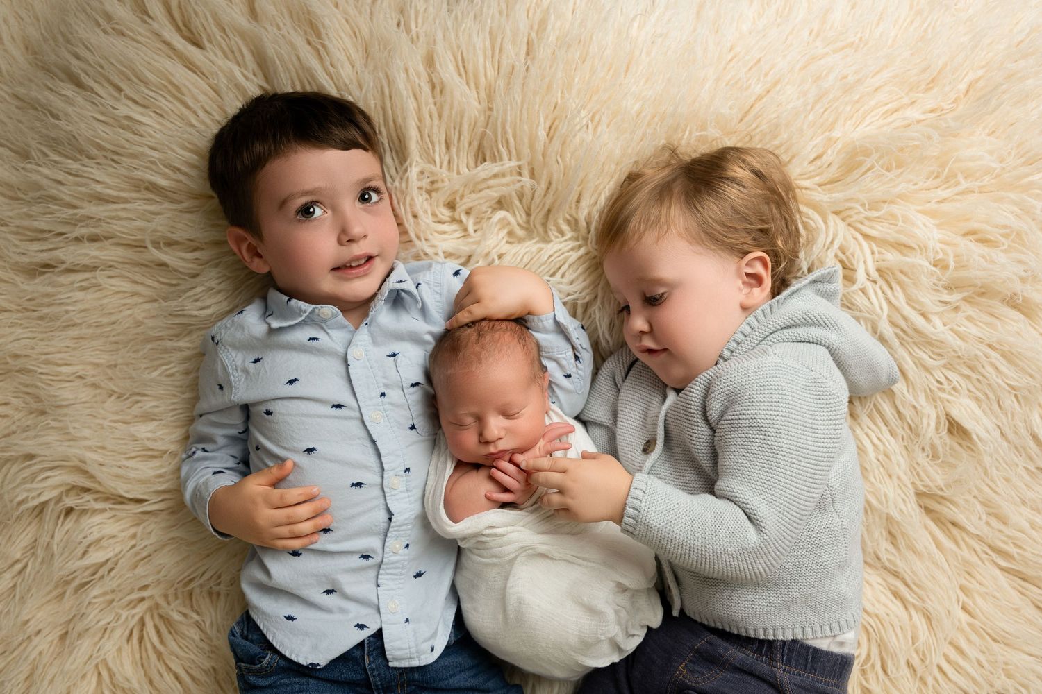 Three siblings lying on fluffy cream sheepskin, with newborn baby in the middle.
