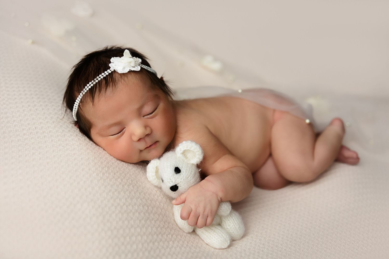 Sleeping newborn cuddling small white teddy bear on textured backdrop with floral headpiece.