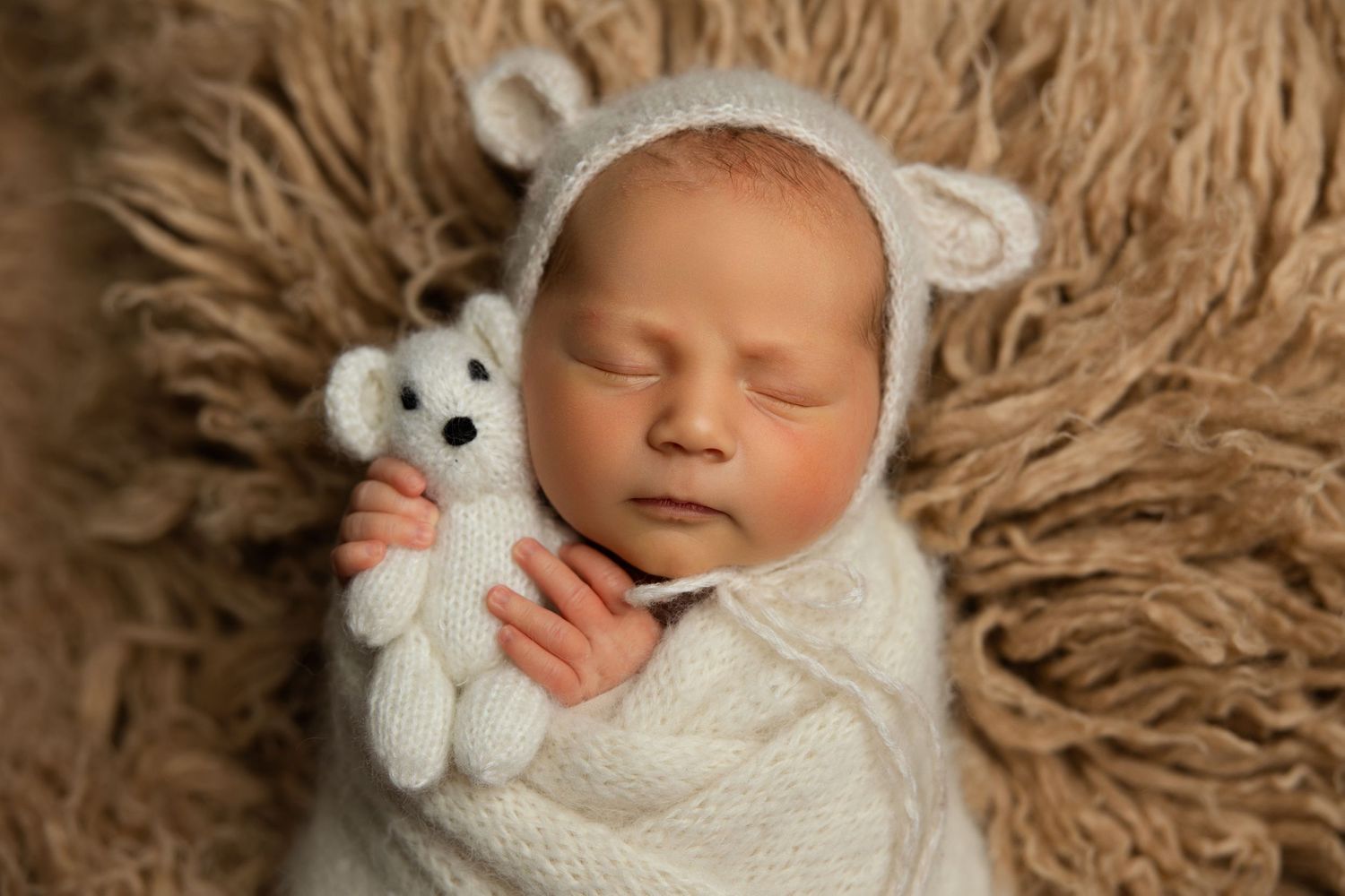 Sleeping newborn wrapped in white swaddle wearing bear ear bonnet with plush toy on furry brown backdrop.