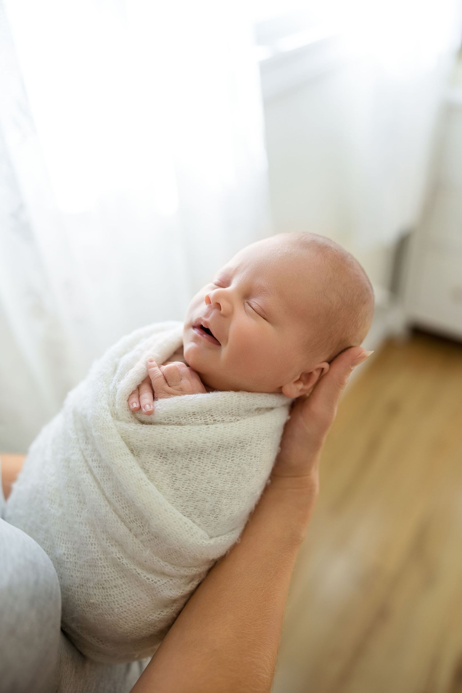 A sleeping newborn baby wrapped in a soft beige blanket is gently cradled in someone's arms.