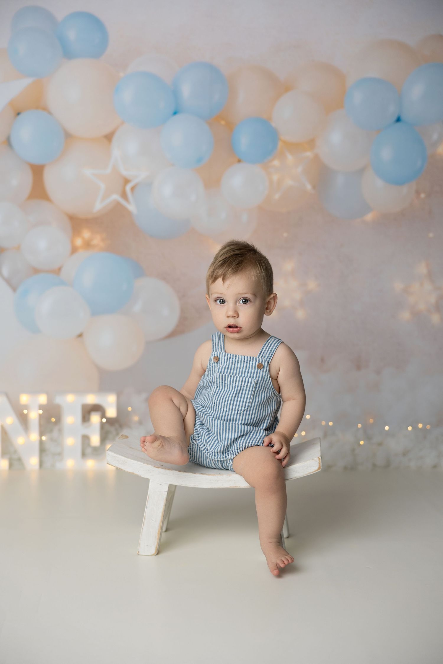 Baby in blue striped romper sitting on white bench with balloon decor and twinkle lights in background.
