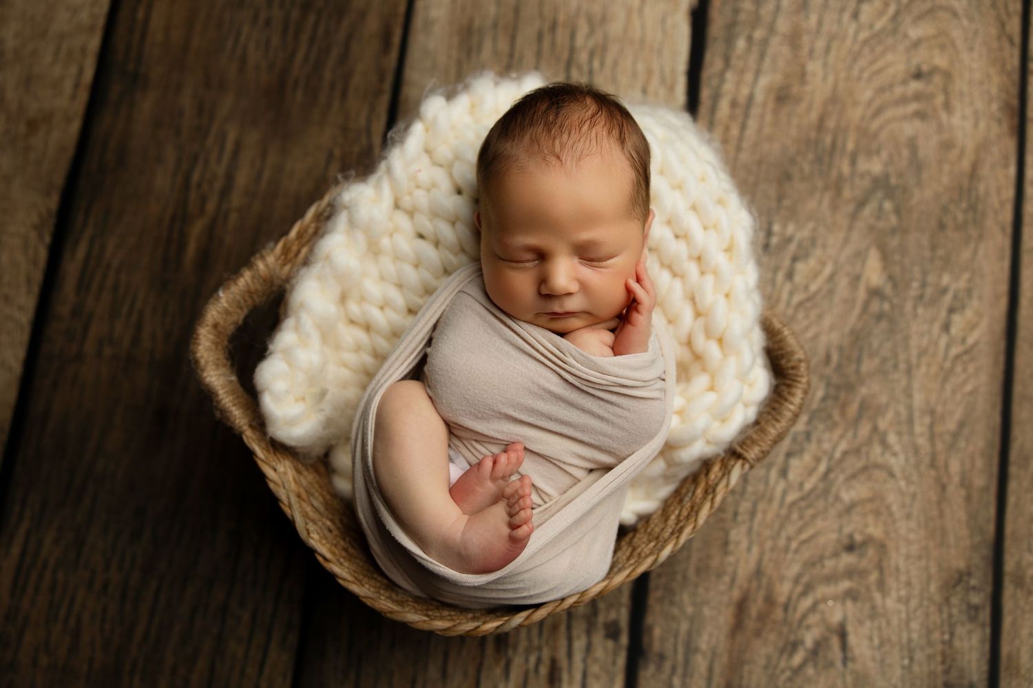 Sleeping newborn wrapped in beige swaddle resting in woven basket.
