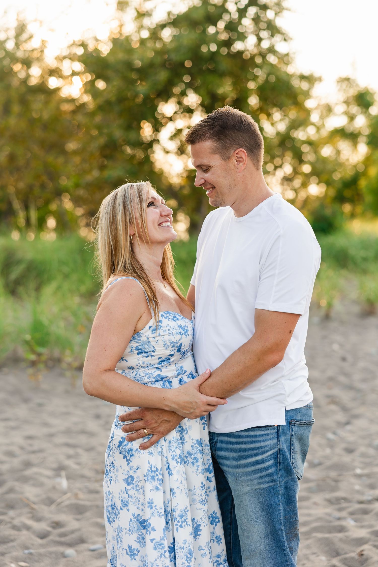 A couple shares an intimate moment on a beach during golden hour with trees in background.