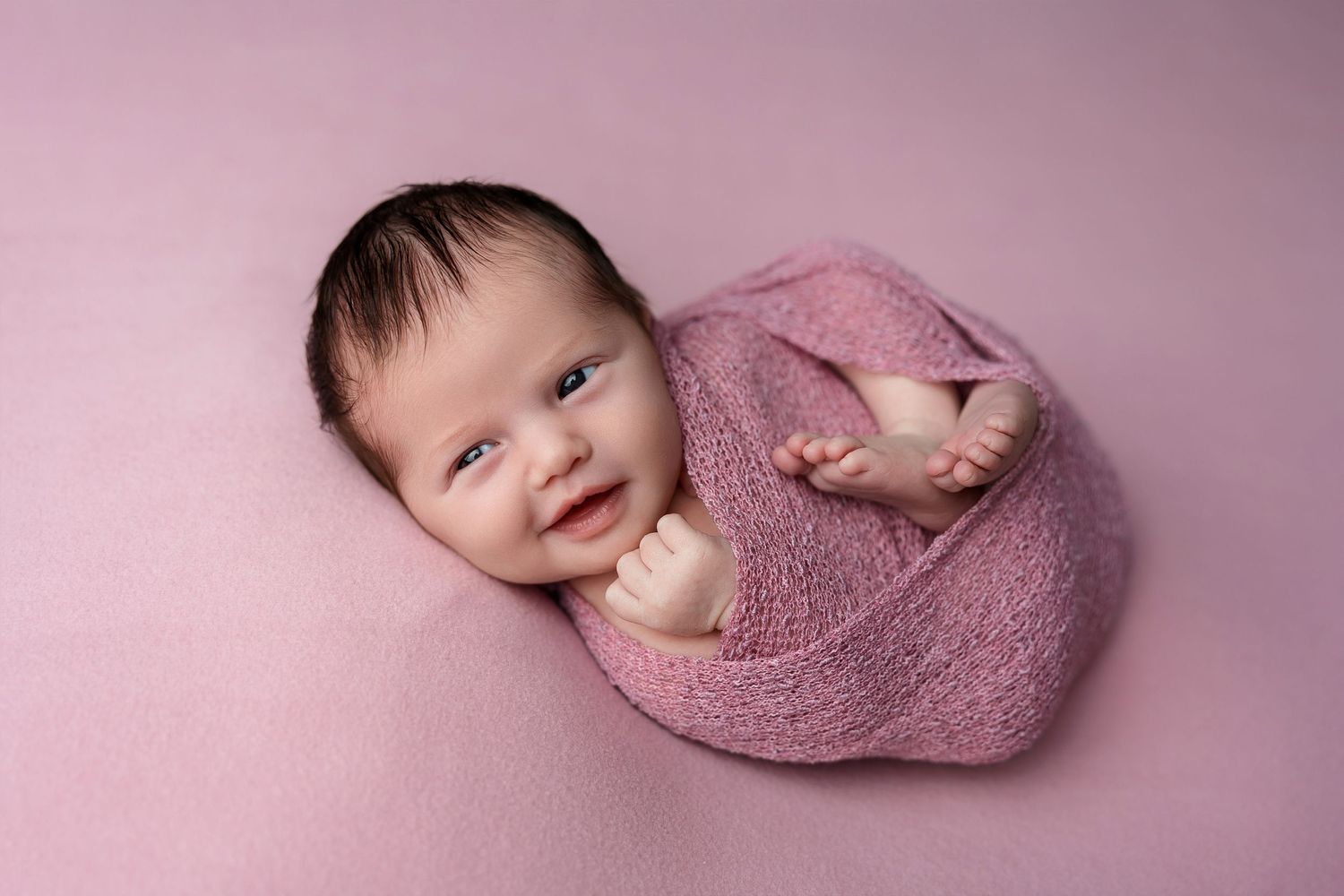 A newborn swaddled in pink fabric displays a sweet smile on a pink background.