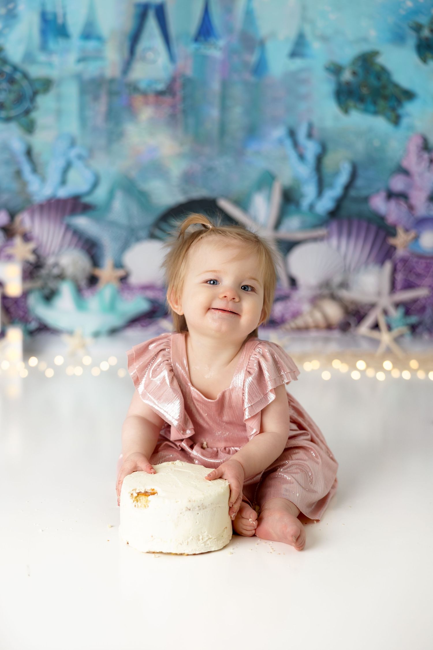 Baby in pink ruffled dress poses with white cake against mystical underwater backdrop.