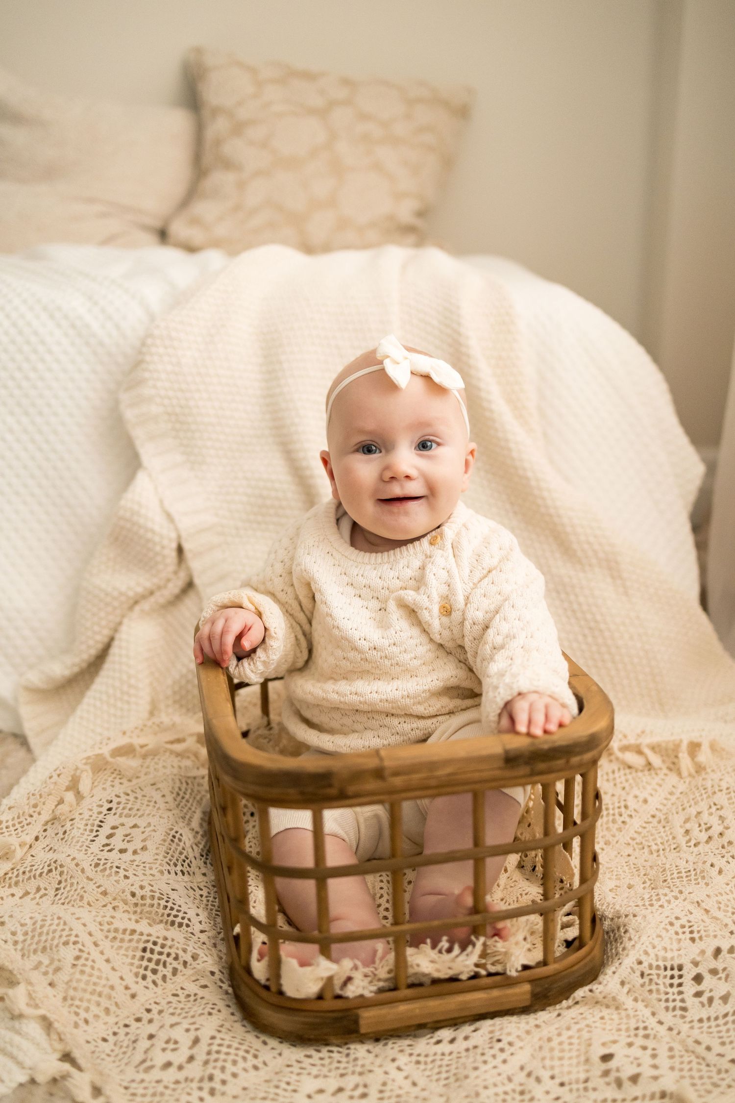 A small baby wearing a cream knit sweater sits inside a vintage wooden rocking cradle on lace fabric.