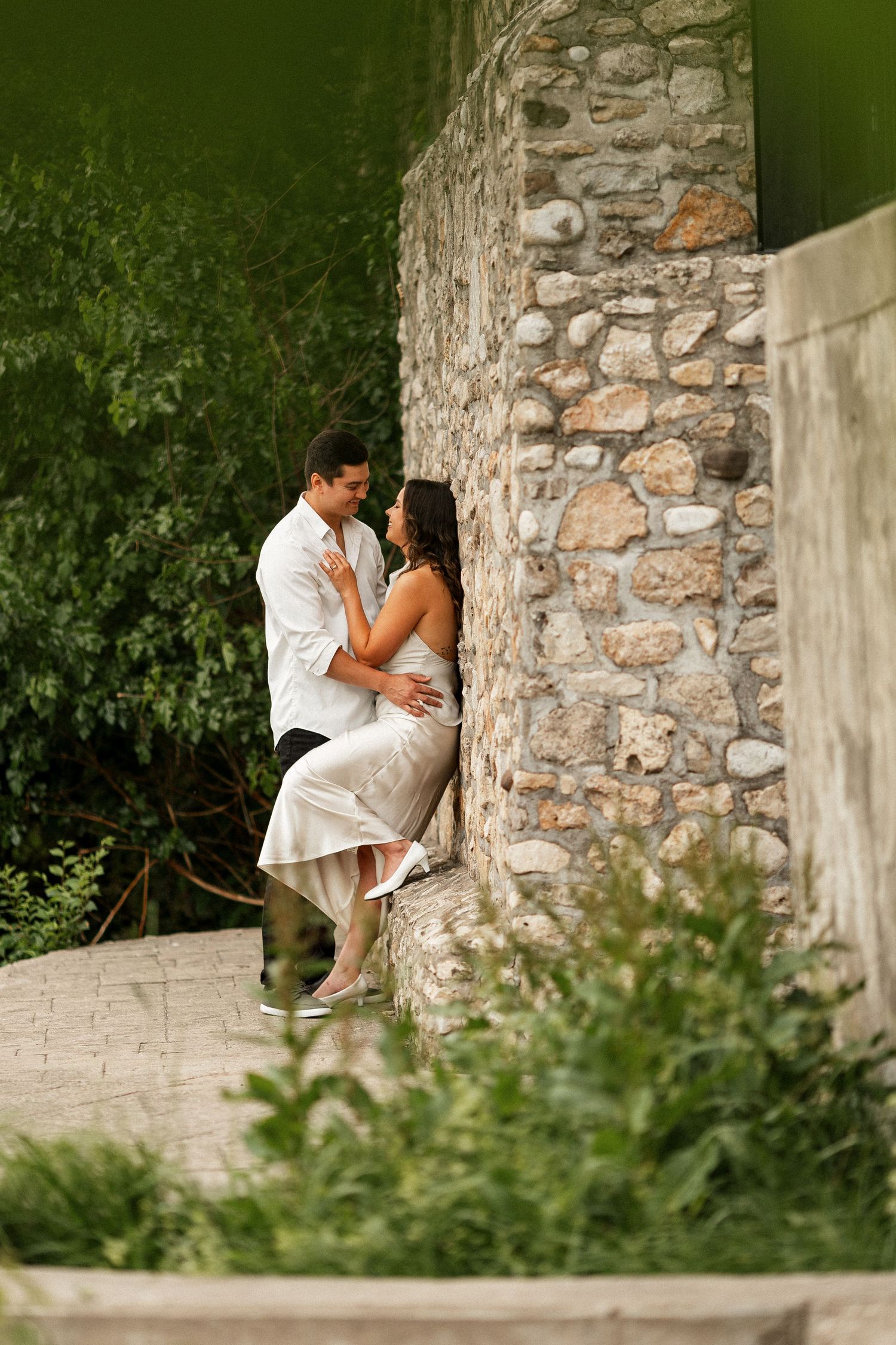 Romantic couple embracing by a rustic stone wall surrounded by lush greenery outdoors.