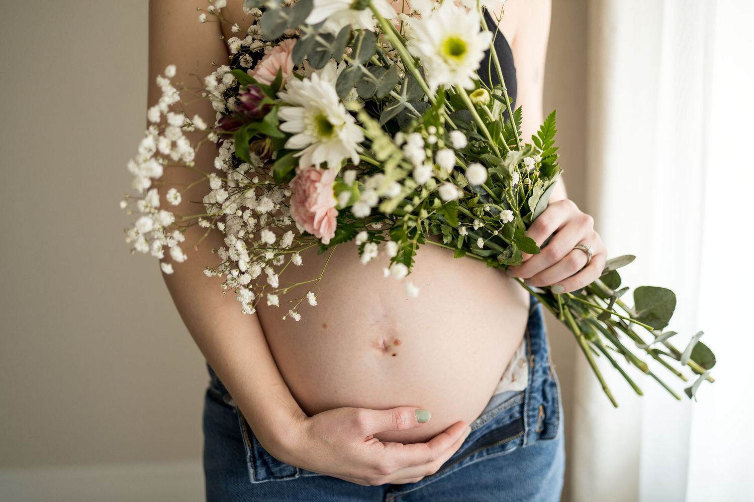 Pregnant woman cradling her bare baby bump while holding a bouquet of white daisies and pink flowers.