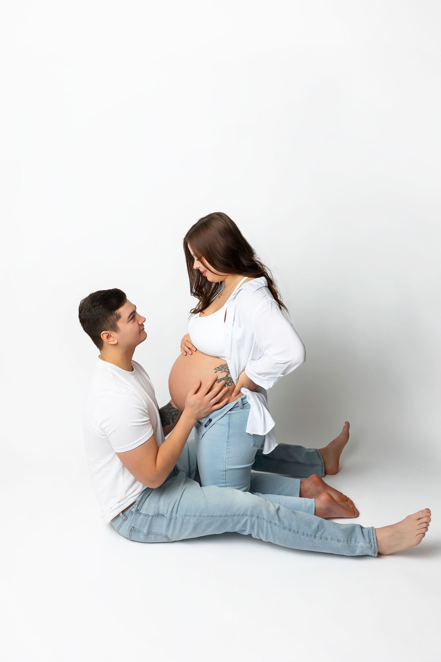 Maternity photo session of a couple in white shirts and jeans against a white backdrop.