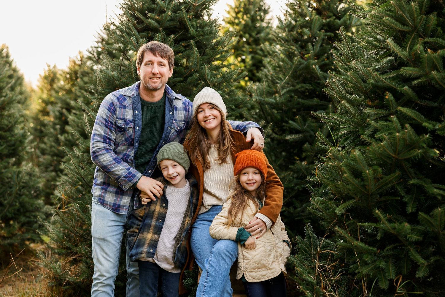 A family stands together smiling among tall evergreen trees at a Christmas tree farm during winter.