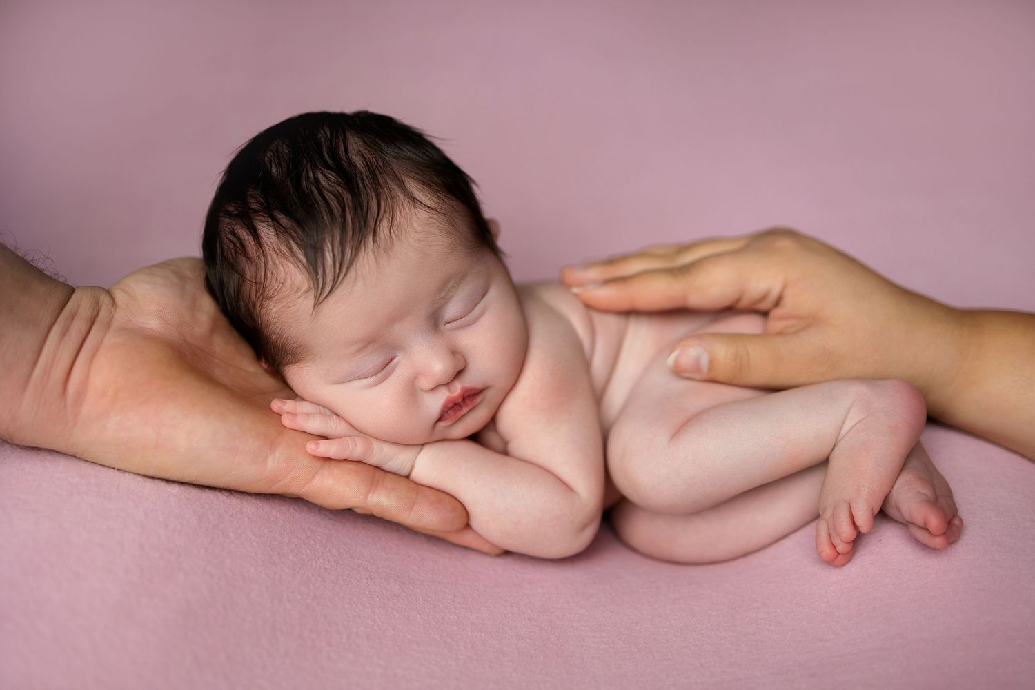 A sleeping newborn rests gently on supporting hands against a pink backdrop.