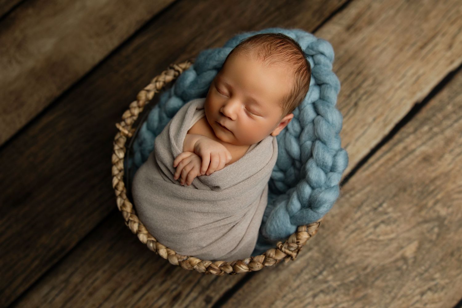 Newborn baby wrapped in gray swaddle sleeping in basket with blue chunky knit blanket on wooden floor.