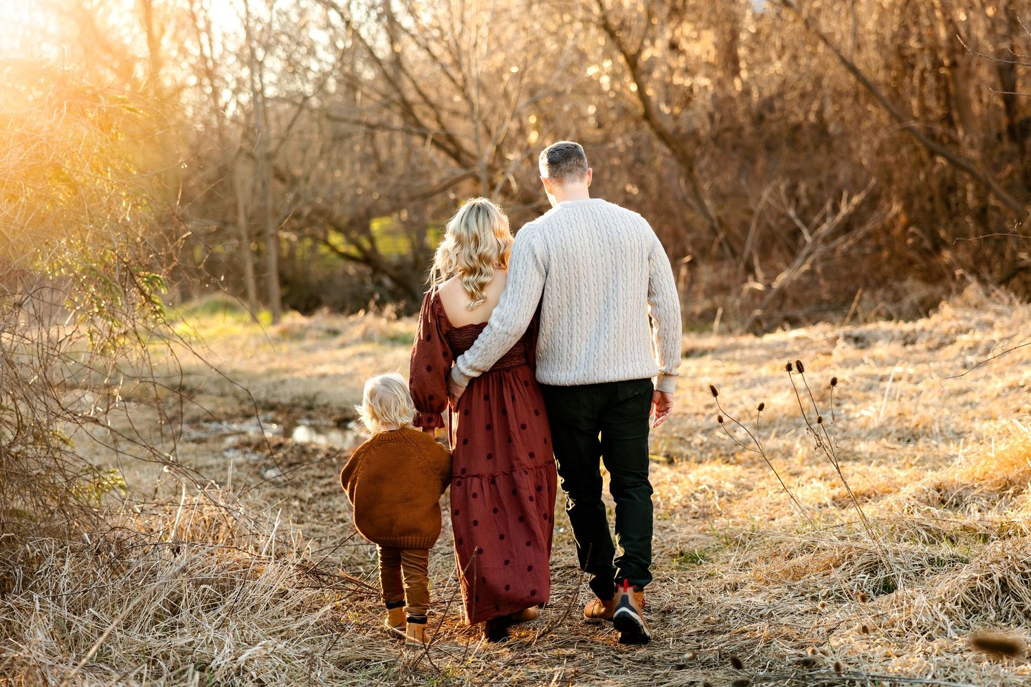 A couple walks hand in hand along a sunlit path through dried winter grass at sunset.