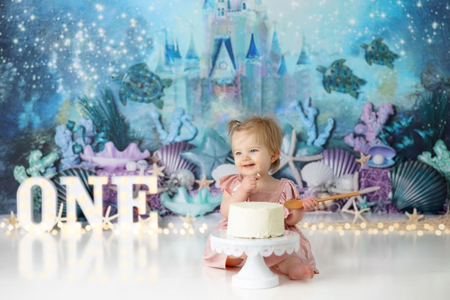 Baby in white outfit explores birthday cake with illuminated ONE sign and underwater themed background.