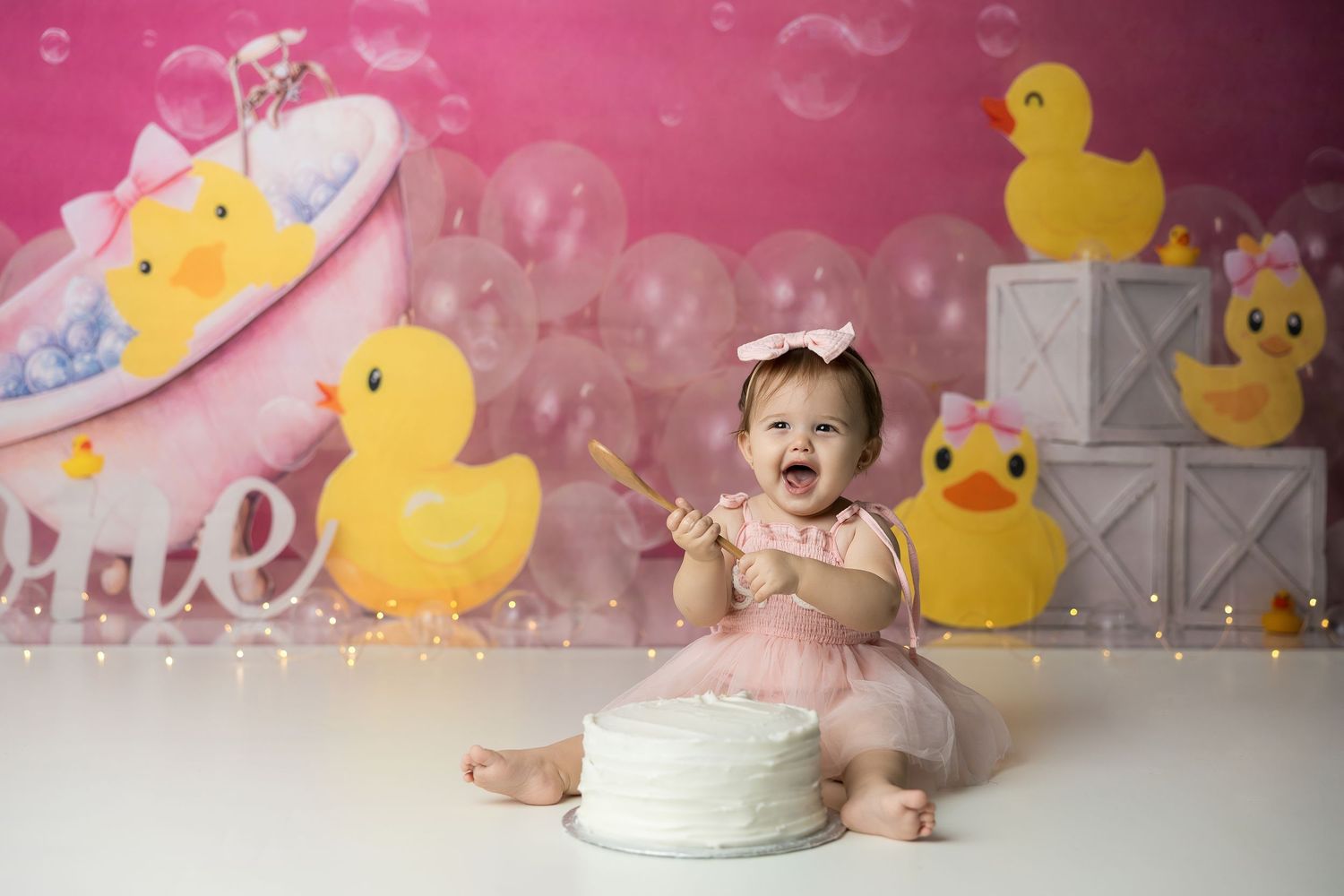 Baby girl in pink tutu smiles at cake smash session with rubber duck themed backdrop and balloons.