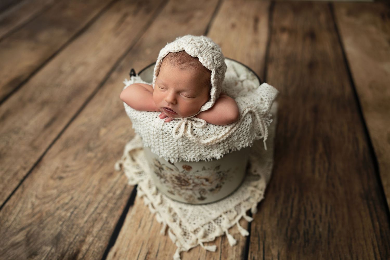 A newborn baby wearing white lace sleeps peacefully in a vintage wooden bowl against a rustic wooden backdrop.