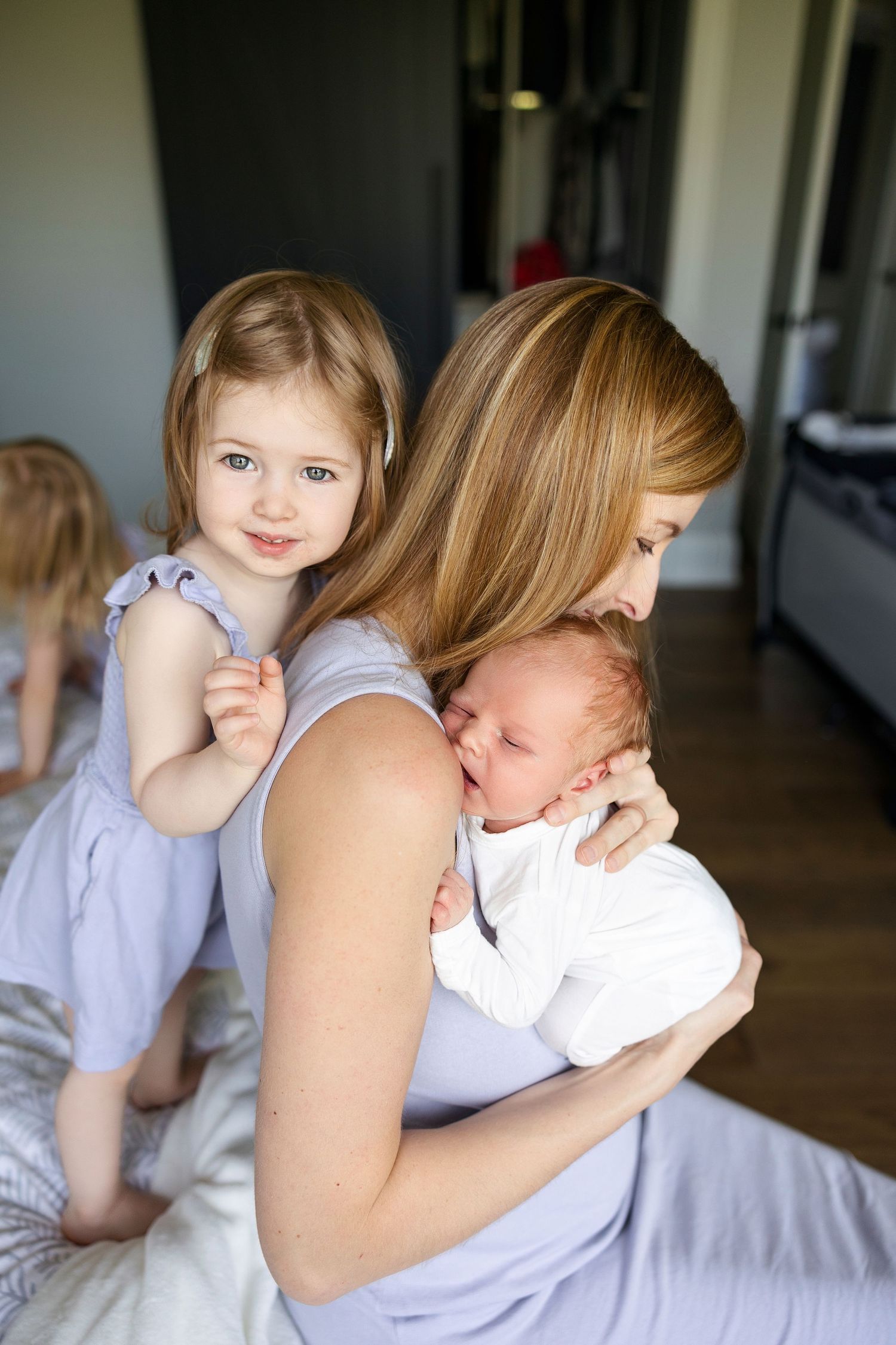A tender family moment captured indoors as a mother in lavender holds a sleeping baby while a toddler stands nearby.