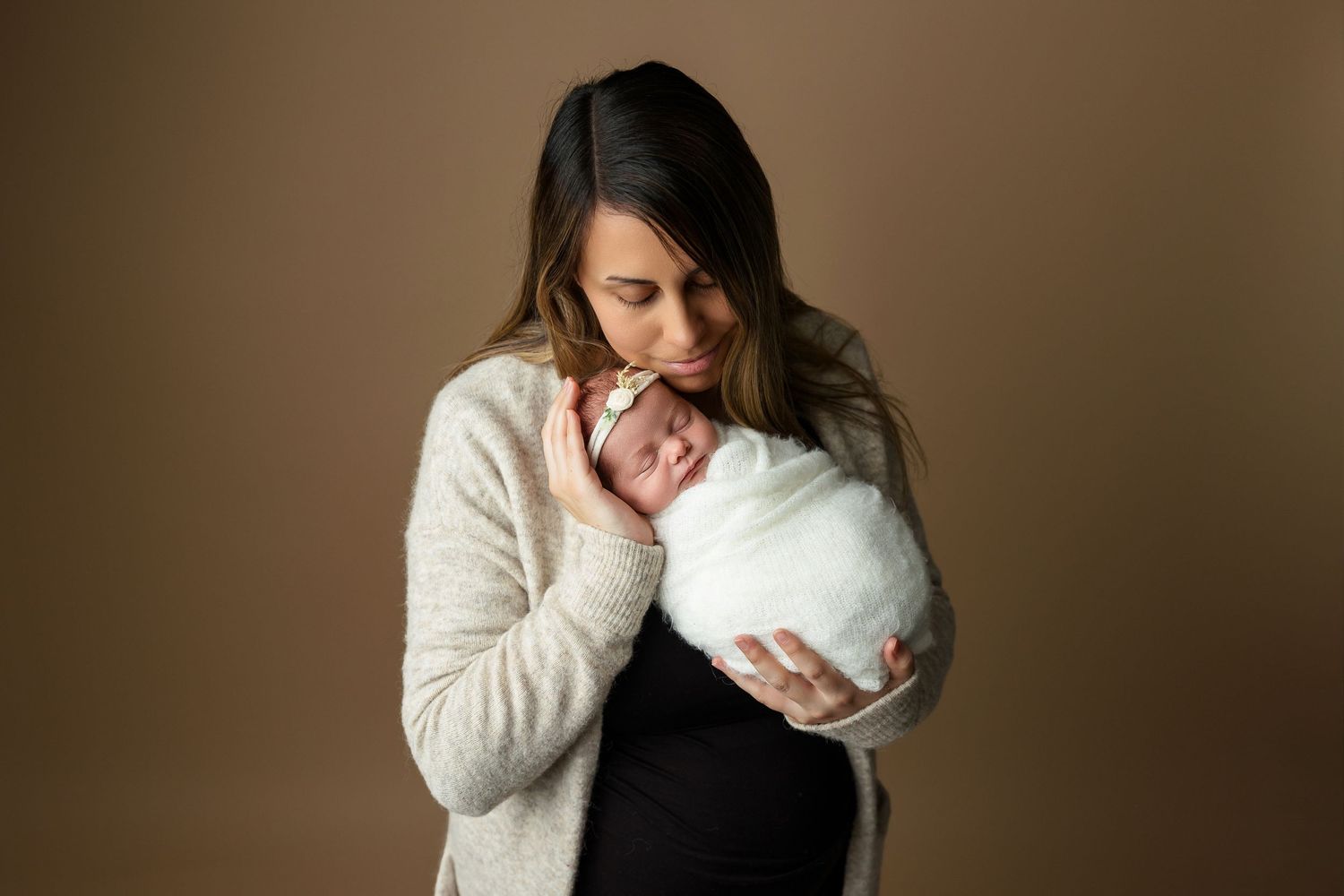 Intimate studio portrait of mother holding swaddled newborn against brown background.