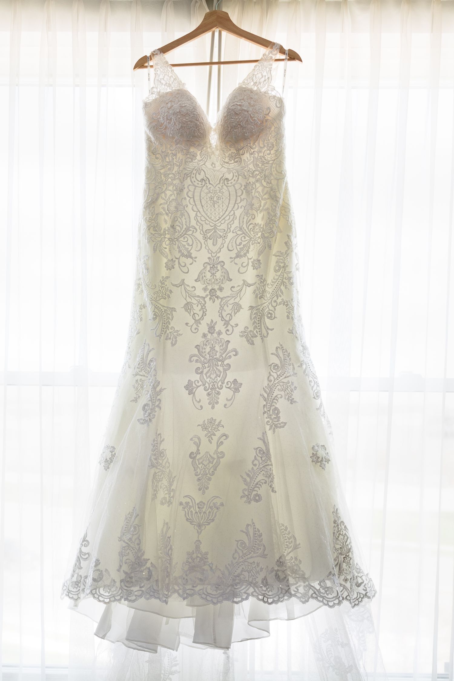An elegant white wedding dress with lace detailing hangs against a light background.