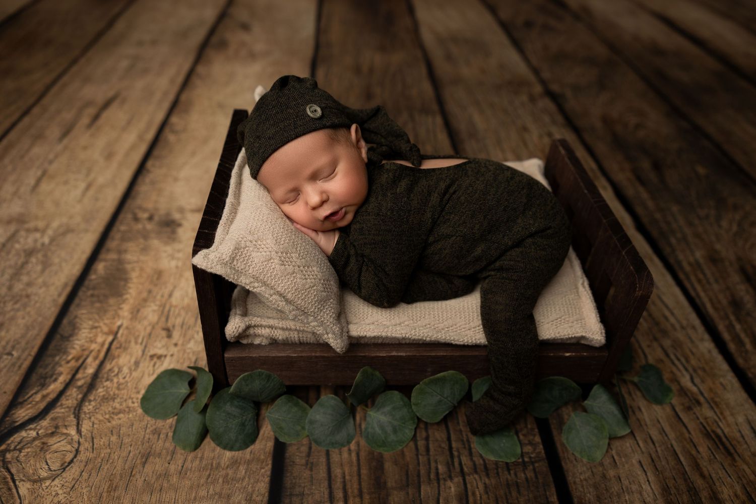 Newborn baby sleeping in black outfit on wooden prop bed with eucalyptus decoration.