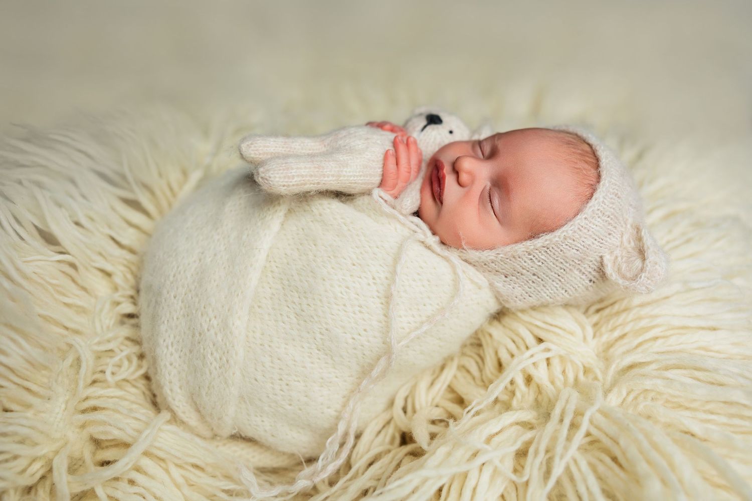 Sleeping newborn with bunny toy wrapped in cream blanket on fluffy white rug.