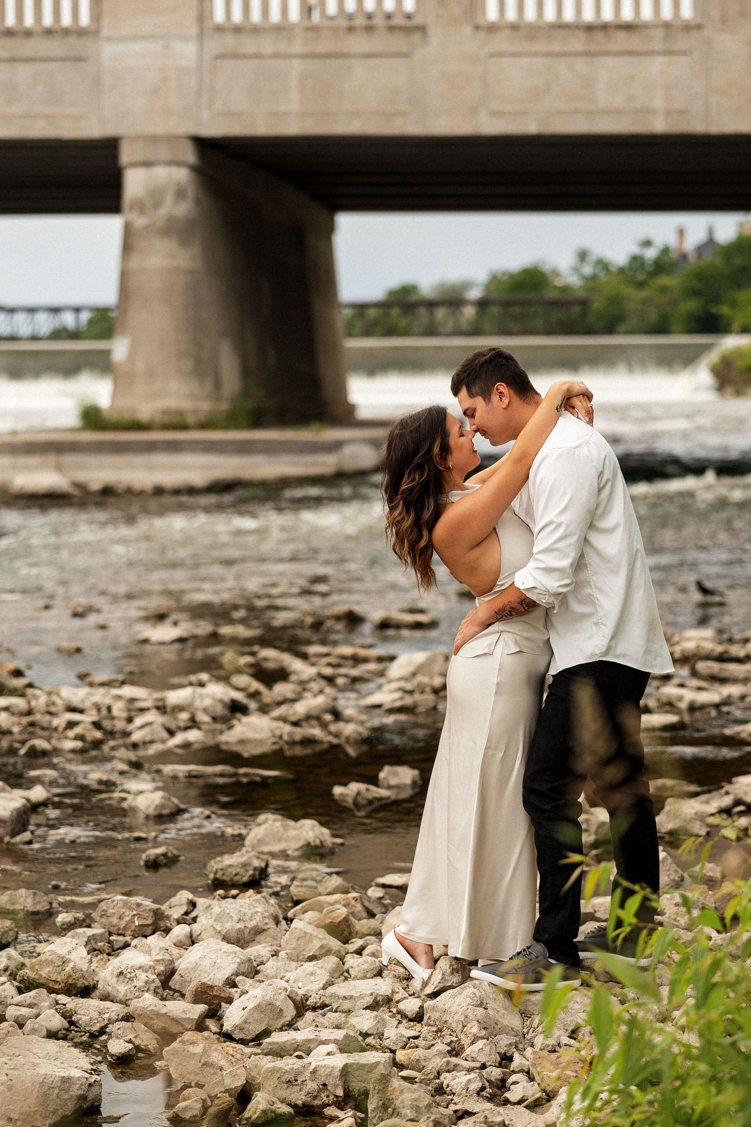 Couple sharing a romantic kiss on rocky riverbank with a concrete bridge and waterfall in the background.