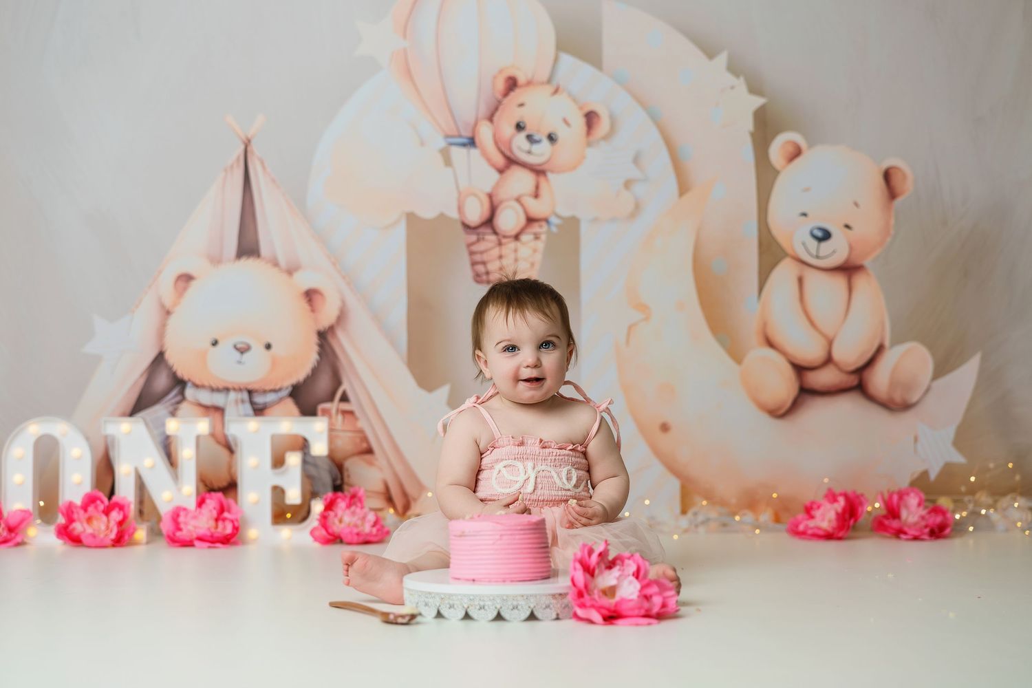 Baby girl first birthday cake smash with pink cake, teddy bear backdrop, and ONE marquee letters.