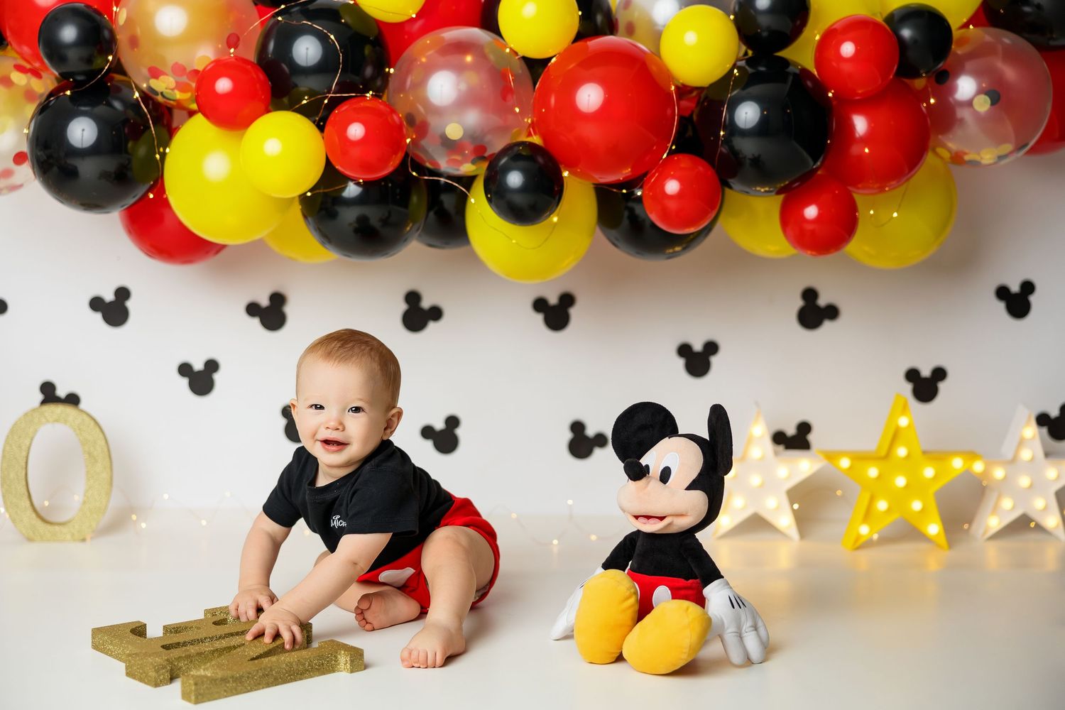 Baby's first birthday Mickey Mouse themed photo with colorful balloons, Mickey plush toy, and festive decorations.