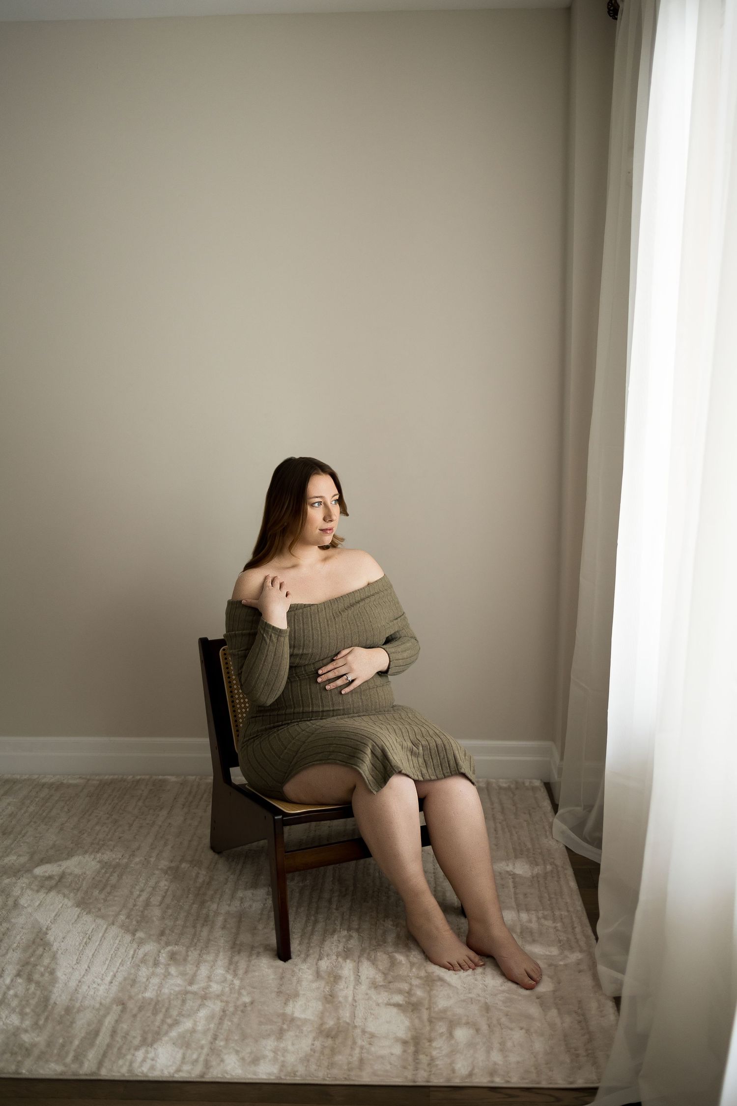 Artistic maternity portraits in olive dress show expectant mother seated in vintage chair by window.