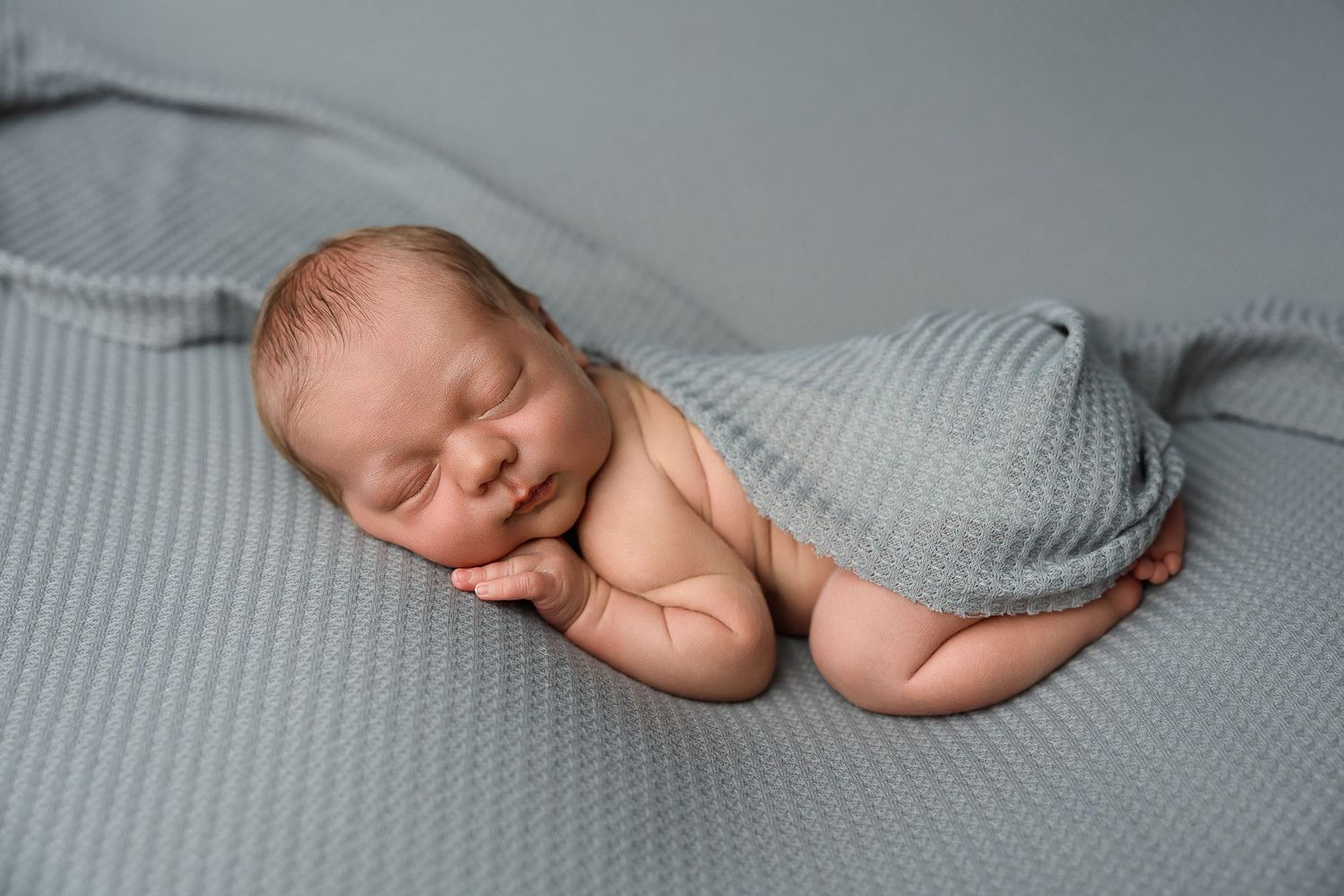 Peaceful newborn baby wrapped in gray knit swaddle sleeping on soft gray blanket.