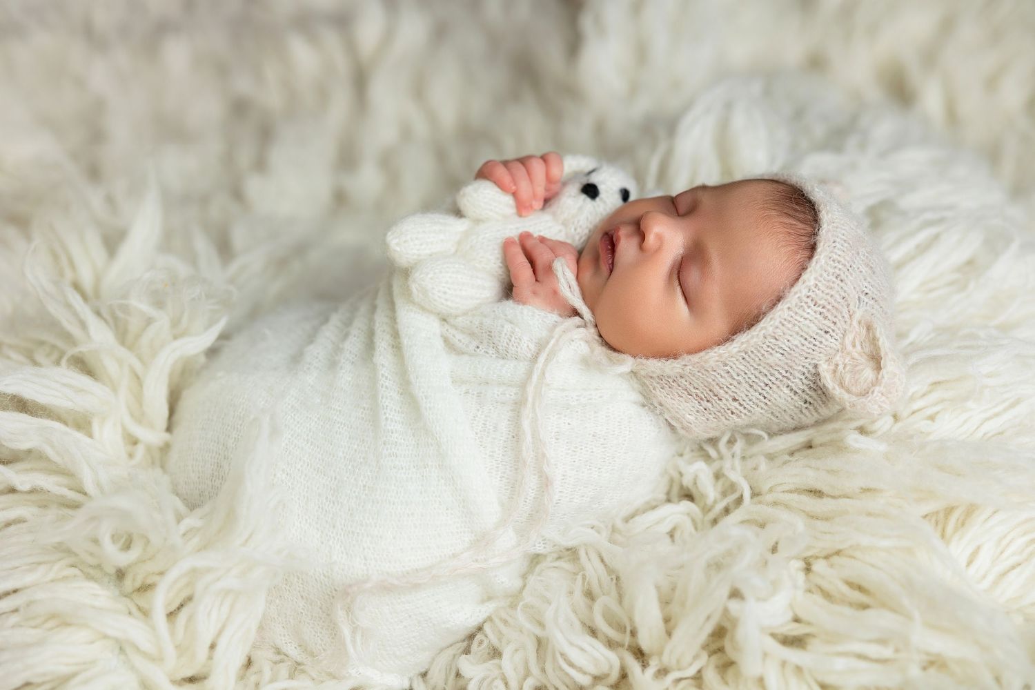 Baby in white outfit lying on fluffy white rug with small stuffed toy.