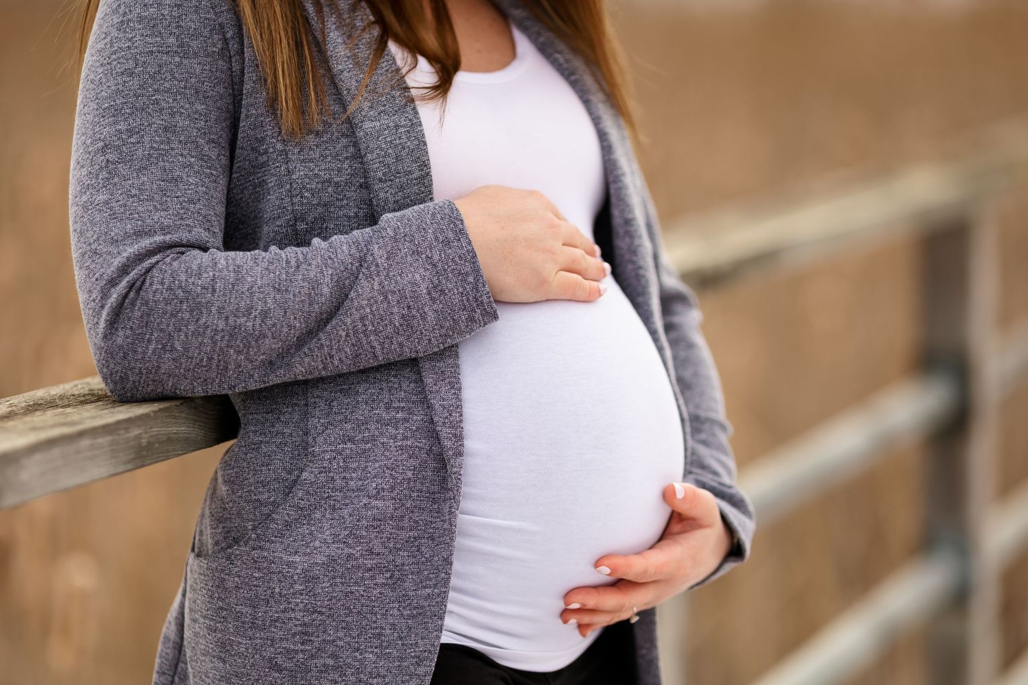 Close up of pregnant belly being cradled in grey cardigan against wooden fence.