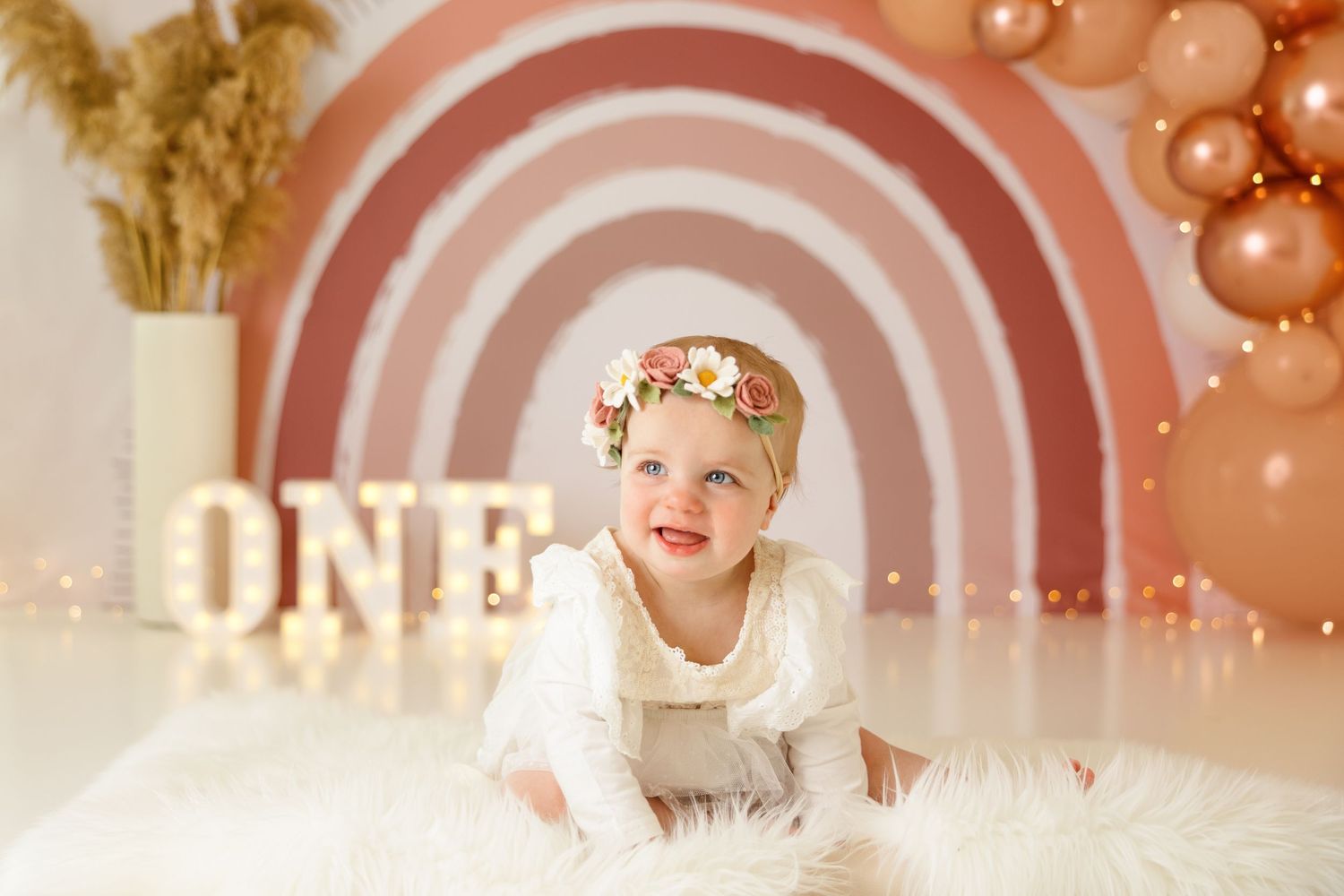 Baby on white fur rug with floral crown against pink rainbow backdrop.